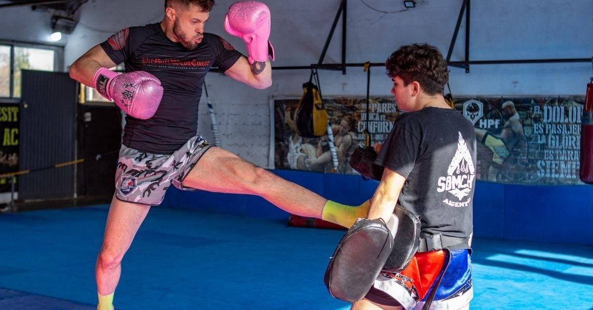 A fighter wearing pink gloves executes a high teep kick into a trainer's pads during a Muay Thai session.