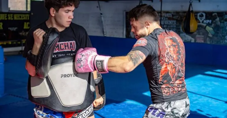 A martial artist in pink boxing gloves throws a punch at a coach wearing a protective body belly pad for Muay Thai training.
