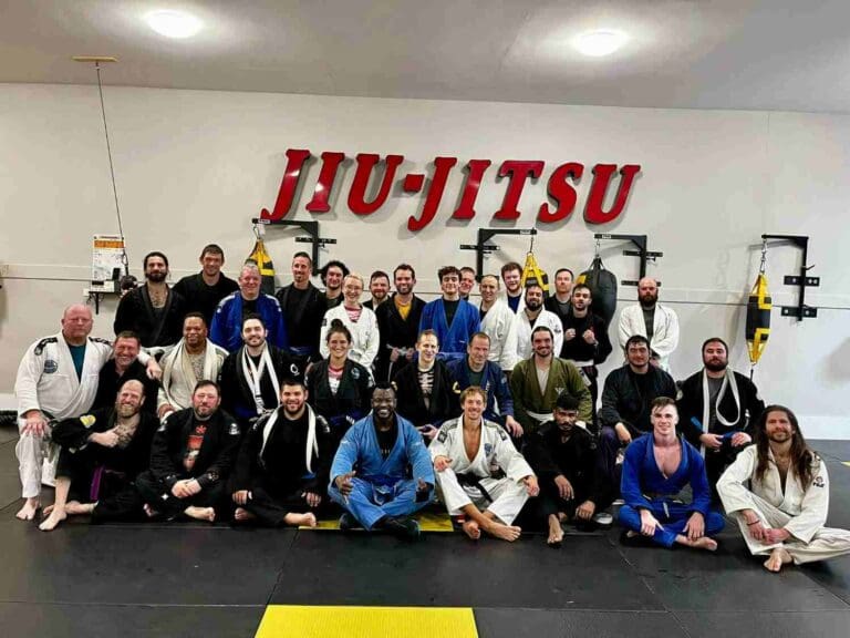 A large group of adult students and coaches sitting and posing together after practice in a martial arts academy, mastering adult Brazilian Jiu Jitsu positional hierarchy.