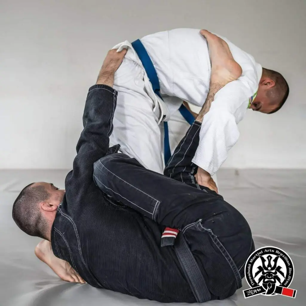 A practitioner in a black gi working an open guard setup against a standing partner during a grappling roll, demonstrating adult Brazilian Jiu Jitsu positional hierarchy.