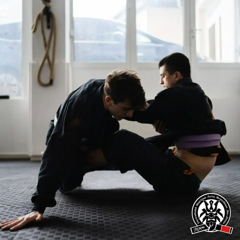 Two adult practitioners engaging in seated guard play and hand fighting during a technical training drill, breaking down adult Brazilian Jiu Jitsu positional hierarchy.