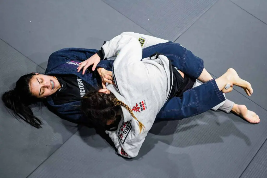 Two female grapplers actively battling in half guard control during a practice session, learning adult Brazilian Jiu Jitsu positional hierarchy.